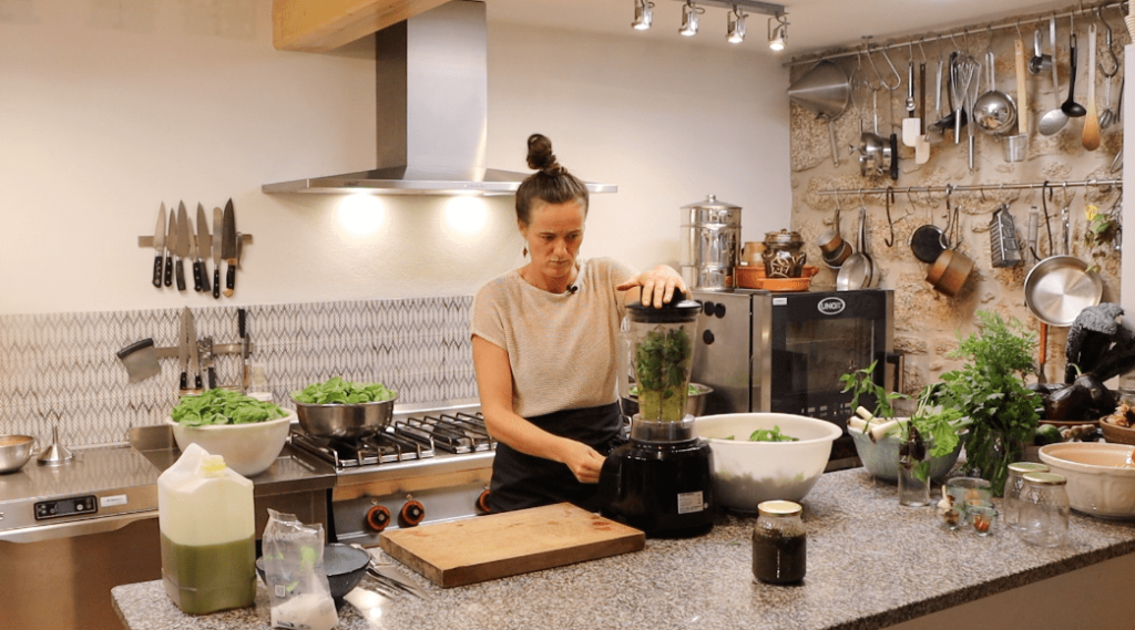 woman using a high-speed blender to blend fresh green basil leaves together with olive oil