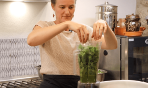 woman putting fresh green basil leaves into a high-speed blender