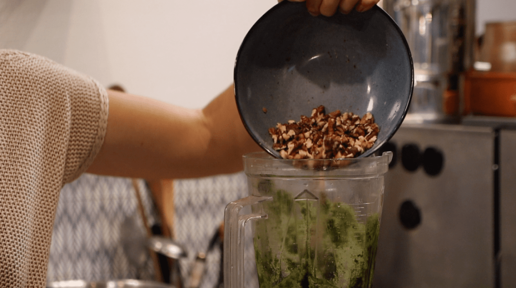 cleaned walnuts dropping from a bowl into a blender with basil and olive oil mixture