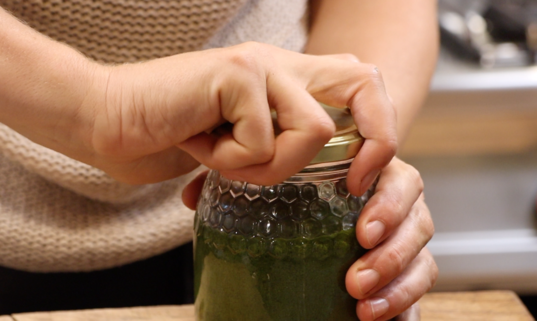 glass jar filled with green paste being closed with a golden metal lid by two hands