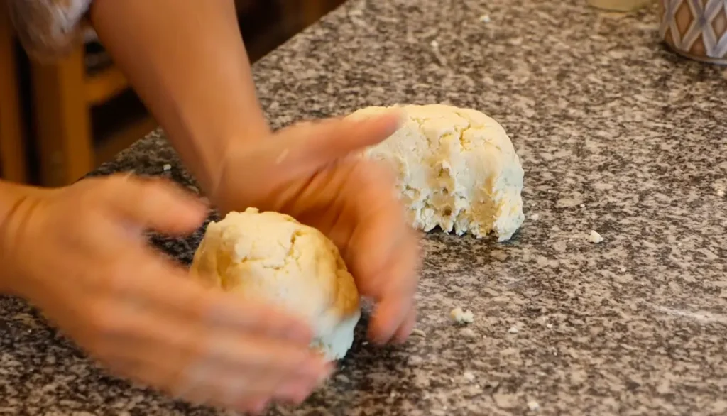 two pieces of dough on stone surface, two hands shaping one into a ball