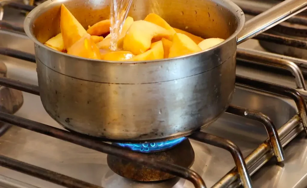 silver pot on stove over blue flame, dark yellow fruit pieces, small stream of water going inside pot