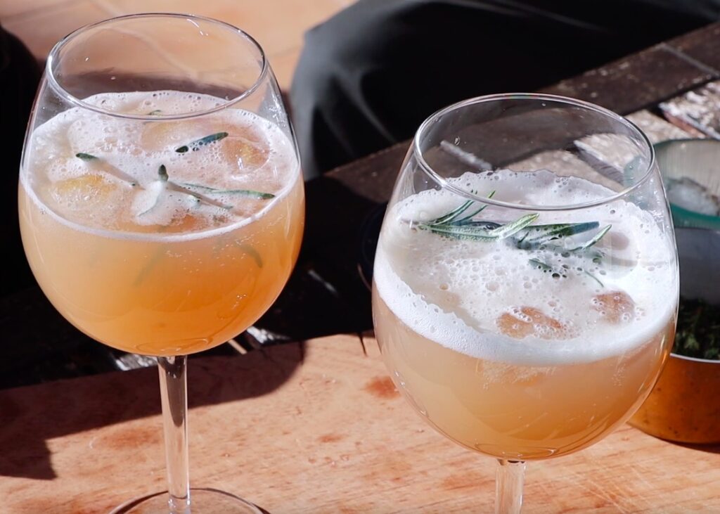 looking down on two wine glasses filled with dark yellow liquid, white foam and branch of herbs