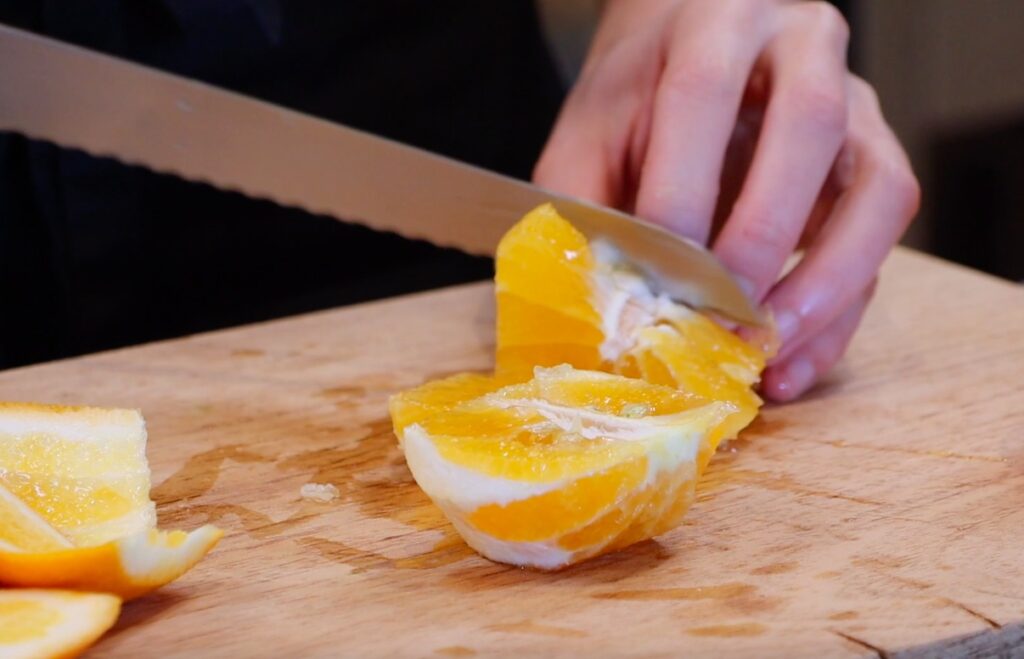 two orange halves being sliced with serrated knife on wooden board