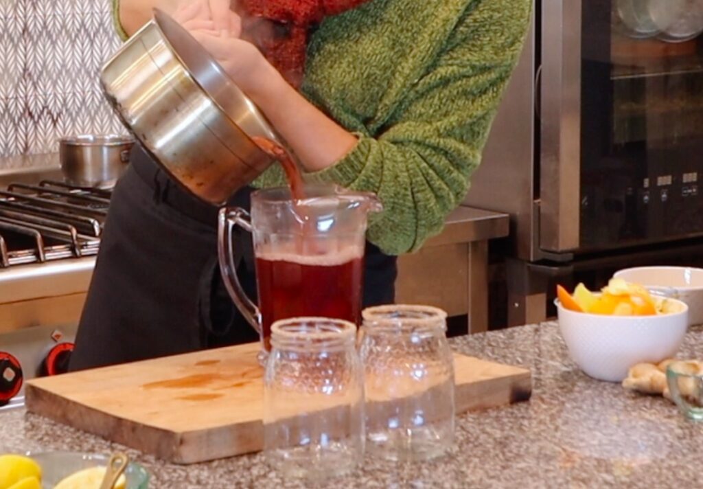 person holding metal pot, tilted, dark red liquid running into glass jug, two jars in front