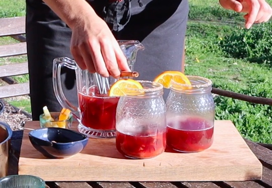 two jars filled one third with dark red liquid, half an orange slice, hand adding cinnamon stick