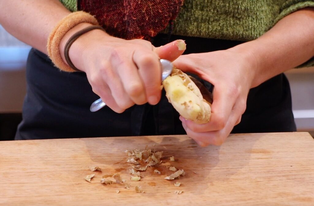 two hands peeling ginger root with a metal spoon