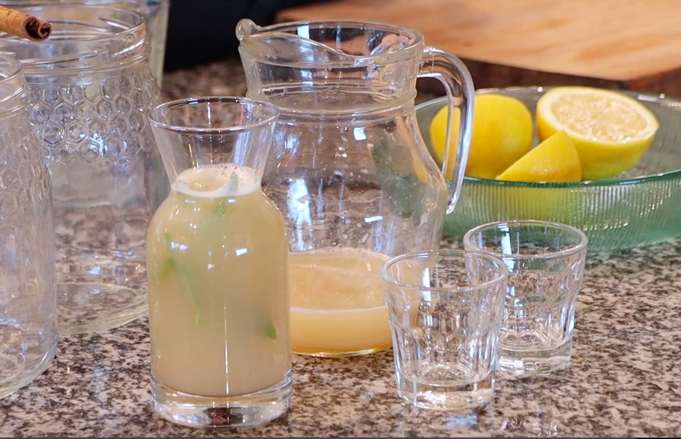 glass jug with yellow liquid, small glass bottle with green leaves and liquid, two shot glasses