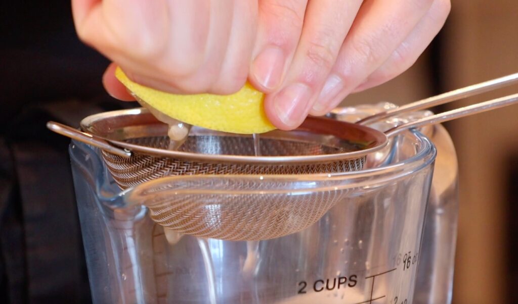 metal sieve on glass measuring cup, two hands squeezing lemon half