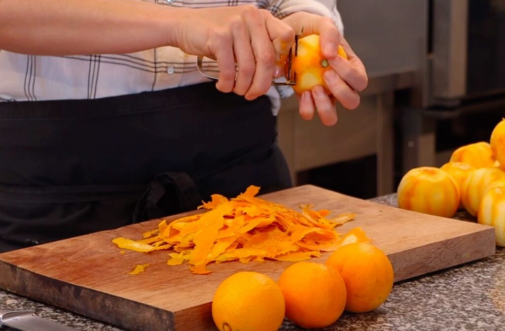 two hands peeling oranges, peels on wooden board, three whole oranges in front