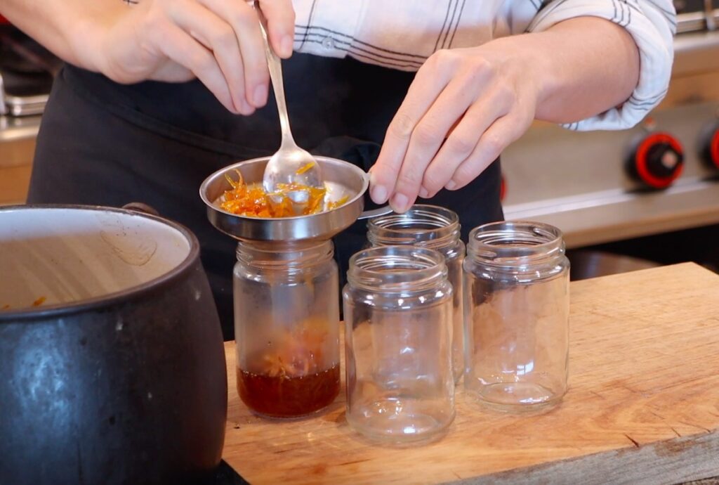 4 glass jars, one with metal funnel, orange marmalade being filled into jars