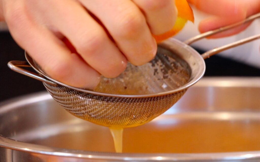 hand squeezing orange, liquid running through metal sieve