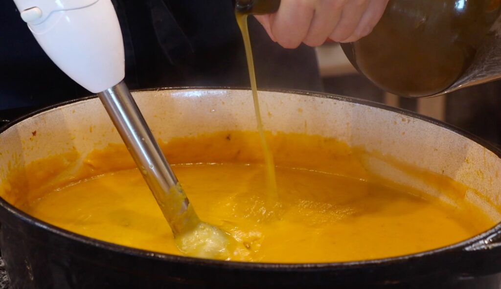 oval pot with smooth puree like orange soup, hand blender, light green liquid being poured