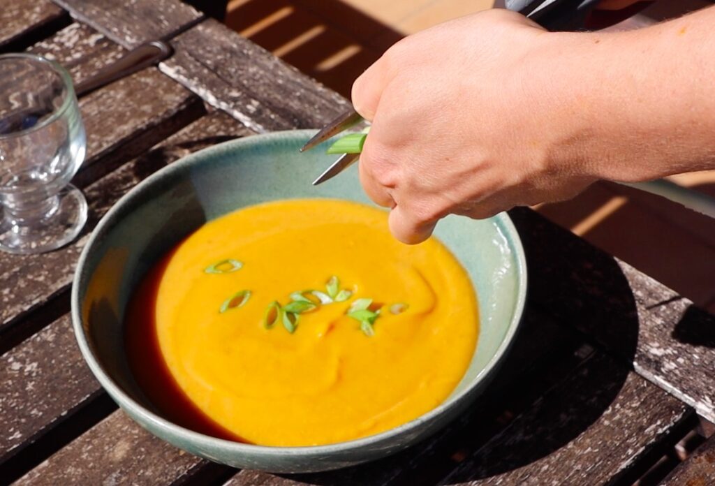 low green soup plate with bright orange soup, hand holding spring onions, scissors, spring onion on soup