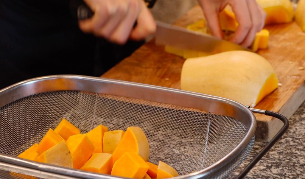 metal sieve with cubes of pumpkin, wooden cutting board, hand with knife and more pumpkin in background